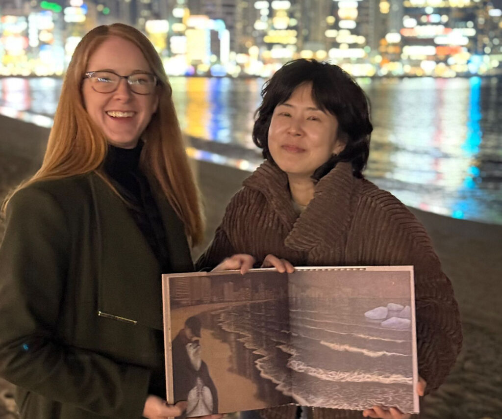 Two women smiling by a beach shoreline at night, holding an open illustrated children’s book featuring a beach scene, with city lights reflecting on the water behind them.