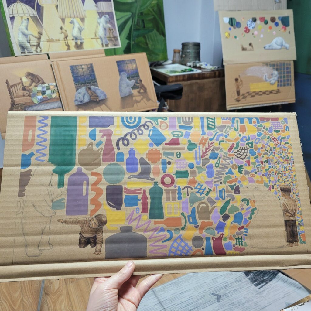 Art studio setup displaying several illustrated children’s book pages with polar bears and children, alongside a colorful mixed media artwork made of various shapes and objects.